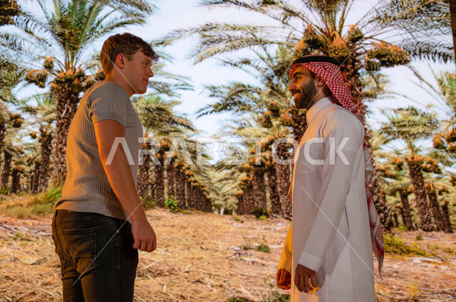 دردشة ومحادثات مع الزوار الاجانب، الدليل السياحي للسياح ،منتجات محليه وطنيه, محاصيل زراعيه سعوديه, موسم حصاد التمور, مزارع النخيل في السعودية ، رجل عربي خليجي سعودي مع سائح أجنبي يرتدي الزي التقليدي يقفان أمام اشجار النخيل المثمرة في محميه طبيعيه