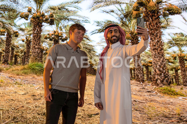 اشجار النخيل المثمرة في موسم حصاد التمور, منتجات محليه وطنيه, محاصيل زراعيه سعوديه, مزارع النخيل في المملكة العربية السعودية ، رجل عربي خليجي سعودي مع سائح أجنبي يقوم بالتقاط صور السيلفي التذكارية عبر الجوال ،محميه طبيعيه لزراعة النخل