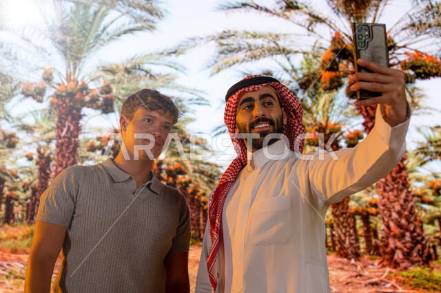 منتجات محليه وطنيه, محاصيل زراعيه سعوديه, اشجار النخيل المثمرة في موسم حصاد التمور, مزارع النخيل في المملكة العربية السعودية ، صورة مقربة لرجل عربي خليجي سعودي مع سائح أجنبي يقوم بالتقاط صور السيلفي التذكارية عبر الجوال ،محميه طبيعيه لزراعة النخل