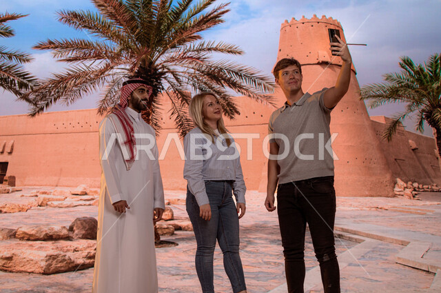 الجدران المتميزه العريضة في البناء المعماري القديم, معالم تاريخية قديمة سياحية, مرشد سياحي عربي ،قصر المصمك التاريخي في مدينة الرياض، شاب عربي سعودي مع السائحين يقوم بتوثيق اللحظات التذكارية بالتقاط صور السيلفي عبر الجوال، حصن المسمك التراثي الطيني