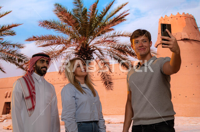 معالم تاريخية قديمة سياحية, مرشد سياحي عربي ،الجدران العريضة المتميزه في البناء المعماري القديم, قصر المصمك التاريخي في مدينة الرياض، شاب عربي سعودي مع السائحين يقوم بتوثيق اللحظات التذكارية بالتقاط صور السيلفي عبر الجوال، حصن المسمك التراثي الطيني