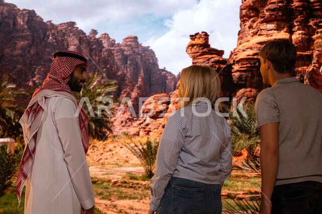 الطبيعه الجبلية المتميزة في المعالم الأثرية التاريخيه السياحيه المشهوره, صورة من الخلف لزائرين اجانب مع مرشد سياحي سعودي يقف في منطقة العلا في السعودية و الاشارة الى شيئ ما , فتح أبواب السياحة السعودية، الدليل السياحي للسياح، وجهه سياحية طبيعيه