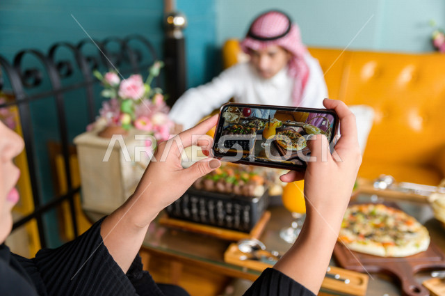 صورة مقربة لسيدة عربية خليجية سعودية تقضى أوقات رائعة في المطعم مع زوجها ، تحمل بيدها الهاتف المحمول و تقوم بإلتقاط صورة خاصة لمائدة الطعام في المطعم ، تشكيلة متنوعة من أصناف الطعام و الماكولات الشهية على الطاولة ، جلسات فريدة و مميزة ، مطاعم و كافيهات في