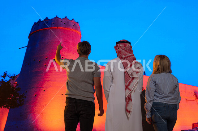 مرشد سياحي عربي ،الجدران العريضة المتميزه في البناء المعماري القديم, متحف توحيد السعوديه, قصر المصمك التاريخي في مدينة الرياض، معالم تاريخية سياحية قديمة, صورة من الخلف لشاب عربي سعودي مع السائحين والاشارة إلى شيء ما، حصن المسمك التراثي الطيني