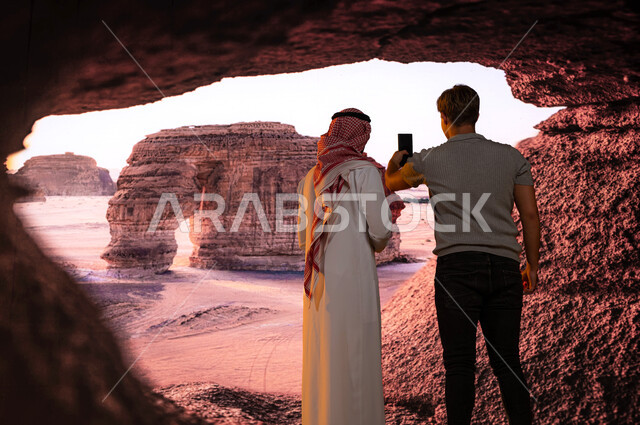 معالم تاريخيه أثرية سياحية في محافظة العلا في السعوديه, فتح أبواب السياحة السعودية، قدوم الزائرين من شتى أنحاء العالم، صورة من الخلف لمرشد سياحي مع سائح أجنبي يلتقط صور تذكارية لجبل الفيل الصخري في الطبيعه الجبلية المميزة في مدينة العلا عبر الجوال
