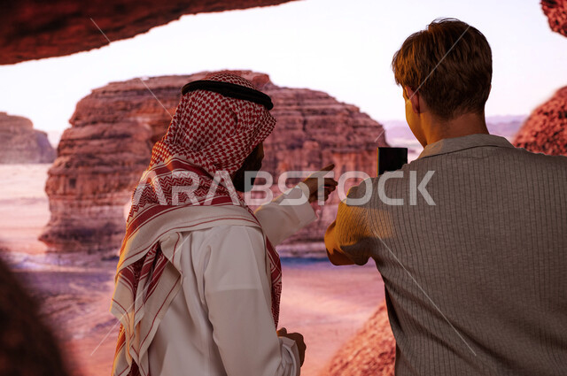 فتح أبواب السياحة السعودية، قدوم الزائرين من شتى أنحاء العالم، معالم سياحيه أثرية في محافظة العلا في السعوديه, صورة مقربة من الخلف لمرشد سياحي مع سائح أجنبي يلتقط صور تذكارية لجبل الفيل الصخري في الطبيعه الجبلية المميزة في مدينة العلا عبر الهاتف المحمول