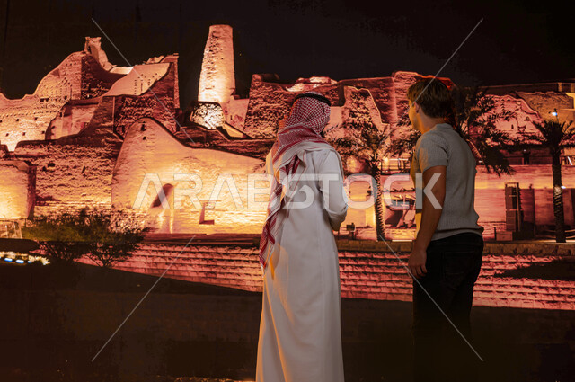 زيارة الآثار والمباني الطينيه في حي البجيري في مدينة الدرعية في الرياض ليلا، منتزه ومعلم تراثي تاريخي على الطراز الشعبي القديم, مركز ثقافي وطني سياحي, صورة من الخلف لمرشد سياحي مع سائح عربي اماراتي خليجي يرتدي العمامة البيضاء والكندورة يقفان امام قصر سلوى