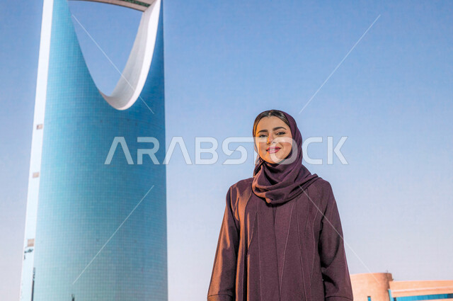 التطور العمراني والنمو , برج المملكة في مدينة الرياض في السعودية ، الفن الهندسي المعماري للابراج وناطحات السحاب، صورة مقربة لإمرأة عربية  خليجية سعودية مبتسمة ترتدي العباءة الخليجية وتنظر الى الكاميرا بإيماءات السعادة، أماكن سياحية مشهورة