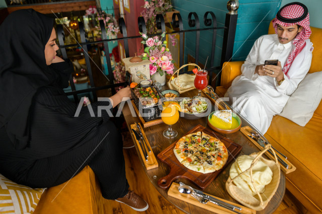 زوجان عربيان خليجيان سعوديان يجلسان على مائدة الطعام و يتناولان الطعام في المطعم ، قضاء اجواء رائعة  في المطعم ، تشكيلة متنوعة من أصناف الطعام و الماكولات الشهية على الطاولة ، جلسات فريدة و مميزة ، مطاعم و كافيهات في السعودية