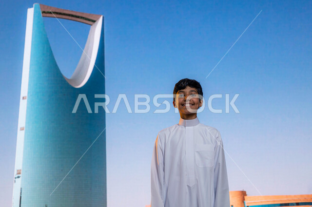 فن الهندسة المعمارية للابراج وناطحات السحاب، معالم سياحية مشهورة, التطور والنمو العمراني, صورة مقربة لولد عربي خليجي سعودي مبتسم يرتدي الثوب التقليدي يقف أمام برج المملكة في مدينة الرياض في السعودية وينظر إلى الكاميرا بإيماءات السعادة