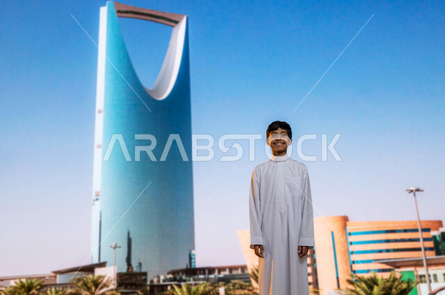الفن الهندسي المعماري للابراج وناطحات السحاب، معالم سياحية مشهورة, التطور والنمو العمراني, ولد عربي خليجي سعودي مبتسم يرتدي  الثوب التقليدي يقف أمام برج المملكة  في مدينة الرياض في المملكة العربية السعودية وينظر إلى الكاميرا