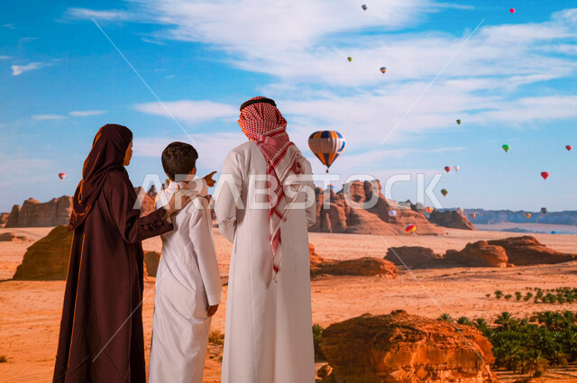 تبادل الأحاديث أمام المعالم الأثرية التاريخية القديمة، الطيران في بالون الهواء الساخن في محافظة العلا, وجهة عائلية سياحية طبيعيه, انشطه وفعاليات ترفيهيه ،صورة من الخلف لزوجان عربيان خليجيان سعوديان مع ولدهما يشير إلى شيء ما، المناطيد في سماء السعودية