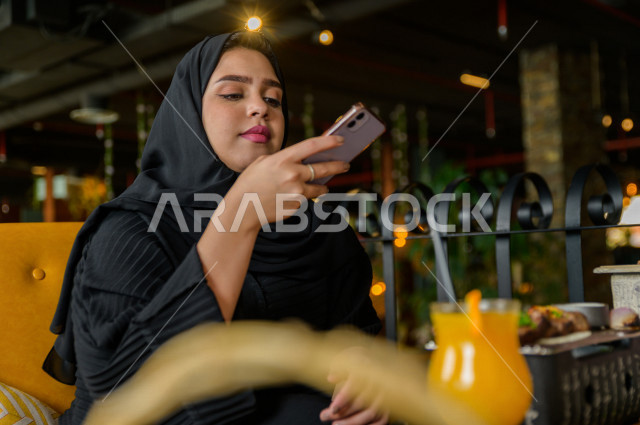 سيدة عربية خليجية سعودية تحمل بيدها الهاتف المحمول و تقوم بإلتقاط صورة خاصة لمائدة الطعام في المطعم ، تشكيلة متنوعة من أصناف الطعام و الماكولات الشهية على الطاولة ، قضاء اجواء رائعة ، جلسات فريدة و مميزة ، مطاعم و كافيهات في السعودية