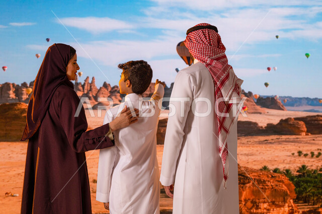 تبادل النقاشات أمام المعالم الأثرية التاريخية القديمة، الطيران في بالون الهواء الساخن في محافظة العلا, وجهة عائلية سياحية طبيعيه, انشطه وفعاليات ترفيهيه ،صورة من الخلف لزوجان عربيان خليجيان سعوديان مع ولدهما يشير إلى شيء ما، المناطيد في سماء السعودية