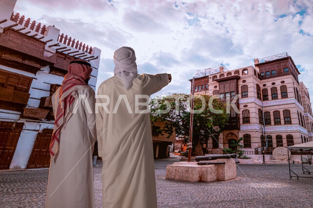 أماكن أثرية سياحية في حي البلد, الإشارة إلى شيء ما، الرواشين الخشبيه في البناء المعماري العربي القديم, صورة من الخلف لسائح عربي اماراتي خليجي يرتدي العمامة والكندورة مع رجل سعودي ينظران إلى المباني الأثرية و الأحياء الشعبيه في جده التاريخيه