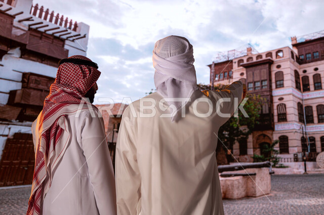 الإشارة إلى شيء ما، الرواشين الخشبيه في البناء المعماري العربي القديم, أماكن سياحيه أثرية في حي البلد, صورة مقربة من الخلف لسائح عربي اماراتي خليجي يرتدي العمامة والكندورة مع رجل سعودي ينظران إلى المباني الأثرية و الأحياء الشعبيه في جده التاريخيه