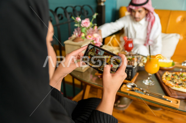 صورة مقربة لسيدة عربية خليجية سعودية تقضى أوقات رائعة في المطعم مع زوجها ، تحمل بيدها الهاتف المحمول و تقوم بإلتقاط صورة خاصة لمائدة الطعام في المطعم ، تشكيلة متنوعة من أصناف الطعام و الماكولات الشهية على الطاولة ، جلسات فريدة و مميزة ، مطاعم و كافيهات في