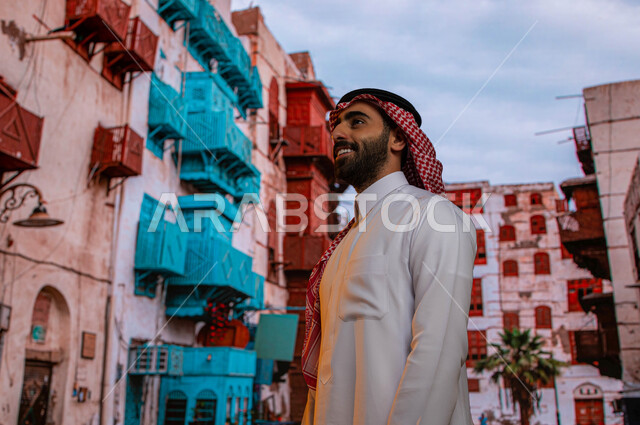 التجول بين الأحياء الشعبيه و المباني الأثرية في جده التاريخيه, صورة مقربة لرجل عربي خليجي سعودي مبتسم يرتدي الثوب والشماغ السعودي ينظر إلى شيء ما, الرواشين الخشبيه في البناء المعماري العربي القديم, أماكن ومعالم سياحيه تاريخيه مشهوره في حي البلد