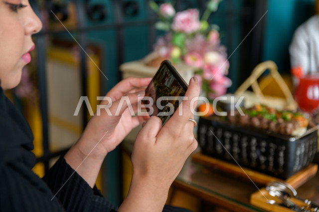 A close-up photo of a Saudi Arabian Gulf woman spending wonderful times in the restaurant with her husband, holding her mobile phone and taking a special picture of the dining table in the restaurant, a variety of food items and delicious foods on the tab