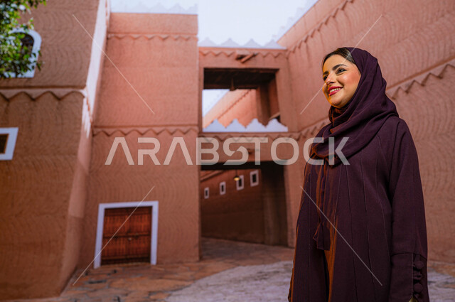 الوقوف في المتحف الوطني السعودي التاريخي في مدينة الرياض، ايماءات تدل على السرور و السعادة, مركز الملك عبد العزيز (قصر المربع) ،معالم تاريخية وطنية سياحية ثقافية في السعودية, امرأه عربية خليجية سعودية مبتسمة محجبة ترتدي العباءه الخليجية تنظر الى شيء ما