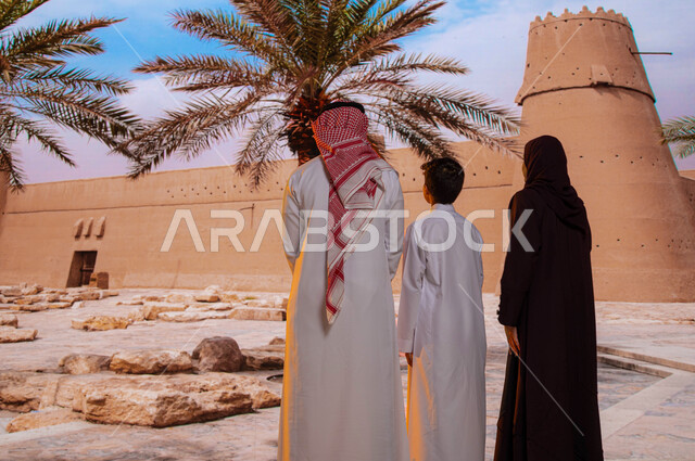 الجدران العريضة المتميزه في البناء المعماري القديم, معالم سياحية تاريخيه قديمة, قصر المصمك التاريخي في مدينة الرياض، صورة من الخلف لعائلة عربية خليجية سعودية تقف أمام متحف توحيد السعوديه و النظر إلى حصن المسمك التراثي الطيني