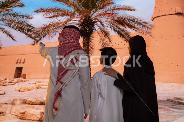 معالم سياحية تاريخيه قديمة, الجدران العريضة المتميزه في البناء المعماري القديم, قصر المصمك التاريخي في مدينة الرياض، صورة من الخلف لعائلة عربية خليجية سعودية تقف أمام متحف توحيد السعوديه, الاشارة الى شيء ما والنظر إلى حصن المسمك التراثي الطيني
