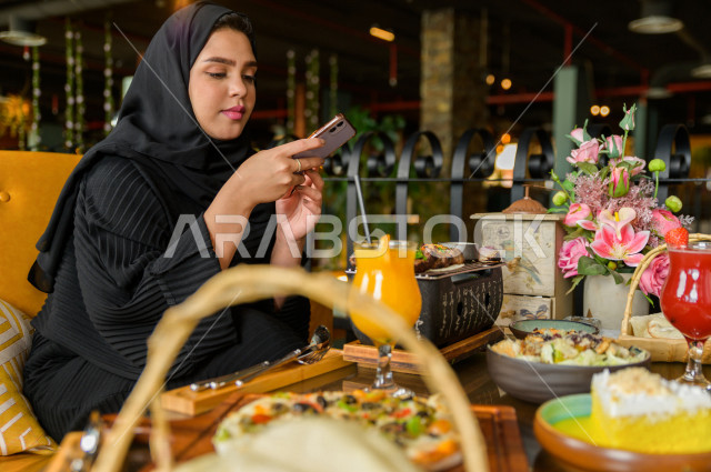سيدة عربية خليجية سعودية تحمل بيدها الهاتف المحمول و تقوم بإلتقاط صورة خاصة لمائدة الطعام في المطعم ، تشكيلة متنوعة من أصناف الطعام و الماكولات الشهية على الطاولة ، قضاء اجواء رائعة ، جلسات فريدة و مميزة ، مطاعم و كافيهات في السعودية