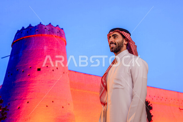 متحف توحيد السعوديه, الجدران المتميزه في البناء المعماري القديم, قصر المصمك التاريخي في مدينة الرياض، معالم تاريخية سياحية قديمة, صورة مقربه لرجل عربي خليجي سعودي مبتسم يرتدي الثوب والشماغ السعودي ويقف أمام حصن المسمك التراثي الطيني