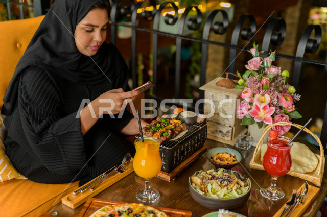 سيدة عربية خليجية سعودية تحمل بيدها الهاتف المحمول و تقوم بإلتقاط صورة خاصة لمائدة الطعام في المطعم ، تشكيلة متنوعة من أصناف الطعام و الماكولات الشهية على الطاولة ، قضاء اجواء رائعة ، جلسات فريدة و مميزة ، مطاعم و كافيهات في السعودية