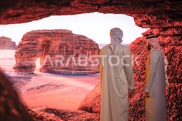 الوقوف أمام جبل الفيل الصخري, معالم تاريخية أثرية سياحيه مشهوره في محافظة العلا في السعوديه, صورة من الخلف لسائح عربي اماراتي خليجي يرتدي العمامة والكندورة الاماراتية مع رجل سعودي يستمتعان بالمناظر الطبيعيه للتكوينات والتشكيلات الصخرية في منطقة العلا .