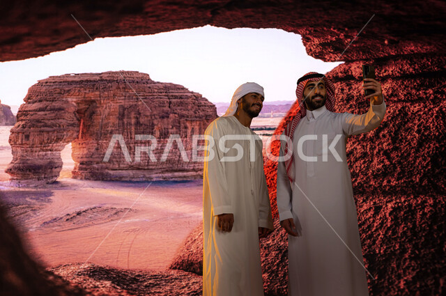 معالم تاريخية أثرية سياحية مشهوره في محافظة العلا في السعوديه, لسائح عربي اماراتي يرتدي العمامة والكندورة مع رجل عربي سعودي يقومان بالتقاط  صور السيلفي عبر الهاتف المحمول أمام جبل الفيل الصخري ،التكوينات والتشكيلات الصخرية في مدينة العلا 