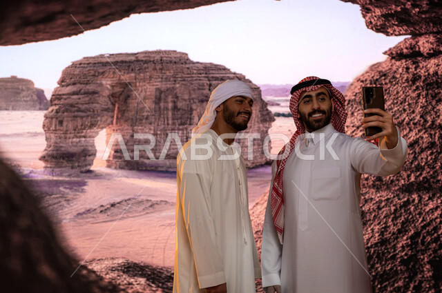 معالم سياحيه تاريخية أثرية  مشهوره في محافظة العلا في السعوديه, صورة مقربة لسائح عربي اماراتي يرتدي العمامة والكندورة مع رجل عربي سعودي يقومان بالتقاط  صور السيلفي عبر الهاتف المحمول أمام جبل الفيل الصخري ،التكوينات والتشكيلات الصخرية في مدينة العلا 