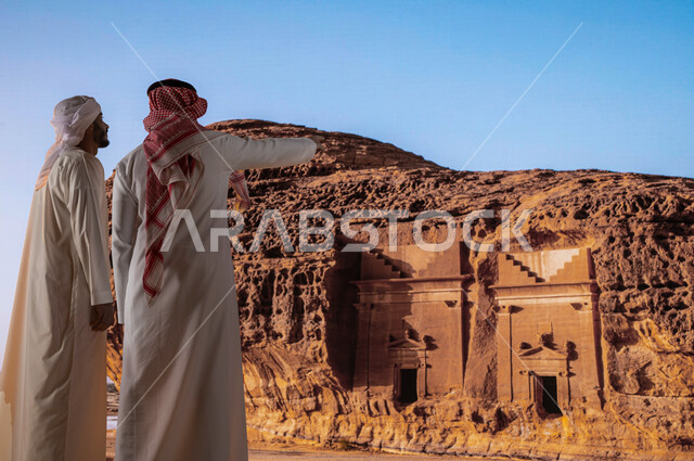 Looking at the formations and rock formations in Madain Saleh in Al-Ula Governorate, tourism in Saudi Arabia, a picture from the back of a Saudi man with an Emirati man wearing a turban and an Emirati kandora enjoying the ancient historical landscapes, th