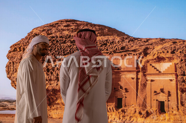 قدوم السياح من شتى أنحاء العالم، النظر إلى التكوينات والتشكيلات الصخرية في مدائن صالح في محافظة العلا، السياحة في السعودية، صورة من الخلف لرجل سعودي مع رجل اماراتي يرتدي العمامة والكندورة الامارتية يستمتع بالمناظر الطبيعيه التاريخية الأثرية القديمة