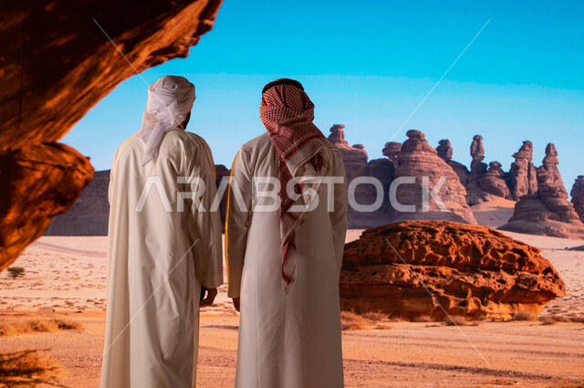 المرتفعات و القمم الجبلية الصخرية ,قدوم السياح من شتى أنحاء العالم، الوقوف أمام التشكيلات و التكوينات الصخرية في محافظة العلا ,صورة من الخلف لرجل سعودي مع سائح عربي اماراتي يرتدي العمامة والكندورة يستمتع بالمناظر الطبيعية الاثرية، السياحة في السعودية