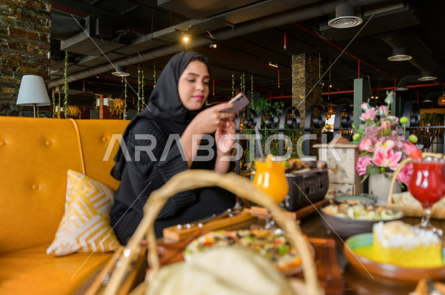 سيدة عربية خليجية سعودية تحمل بيدها الهاتف المحمول و تقوم بإلتقاط صورة خاصة لمائدة الطعام في المطعم ، تشكيلة متنوعة من أصناف الطعام و الماكولات الشهية على الطاولة ، قضاء اجواء رائعة ، جلسات فريدة و مميزة ، مطاعم و كافيهات في السعودية