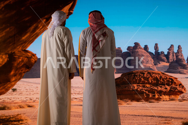 السياحة في السعودية، المرتفعات و القمم الجبلية الصخرية ,قدوم السياح من شتى أنحاء العالم، الوقوف أمام التشكيلات و التكوينات الصخرية في محافظة العلا ,صورة من الخلف لرجل سعودي مع سائح عربي اماراتي يرتدي العمامة والكندورة يستمتع بالمناظر الطبيعية الاثرية