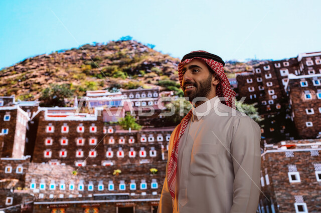 معالم ومناطق سياحيه مشهوره في السعوديه, ايماءات تدل على السعادة، صورة مقربة لرجل عربي خليجي سعودي يرتدي الثوب والشماغ السعودي يقف أمام الآثار و البيوت التاريخية القديمة في أبها ،قرية رجال ألمع التراثية في منطقة عسير