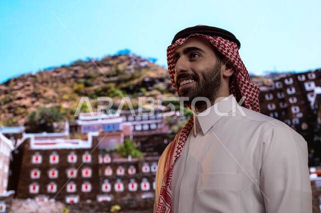 مناطق ومعالم سياحيه مشهوره في السعوديه, ايماءات تدل على السعادة، صورة مقربة لرجل عربي خليجي سعودي يرتدي الثوب والشماغ السعودي يقف أمام قرية رجال ألمع التراثية في منطقة عسير, الآثار و البيوت التاريخية القديمة في أبها