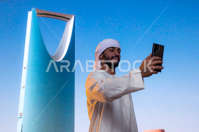 معالم سياحية مشهورة, التطور و النمو العمراني, رجل عربي اماراتي خليجي يرتدي العمامة البيضاء والكندورة الاماراتية يقوم بالتقاط صور السيلفي التذكارية أمام برج المملكة عبر الجوال في مدينة الرياض في السعودية, الفن الهندسي المعماري للابراج وناطحات السحاب