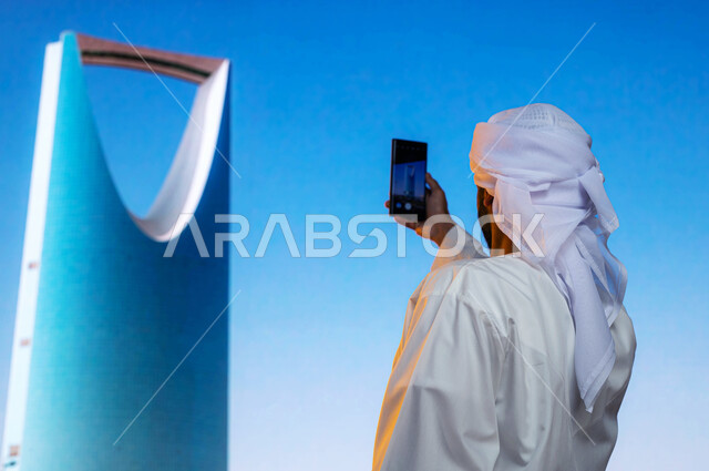 التطور و النمو العمراني, صورة من الخلف لسائح عربي اماراتي خليجي يرتدي الكندورة والعمامة البيضاء الاماراتية يلتقط الصور التذكارية لبرج المملكة في مدينة الرياض في المملكة العربية السعودية, الفن الهندسي المعماري للابراج وناطحات السحاب، معالم سياحية مشهورة