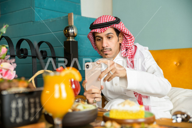 رجل عربي خليجي سعودي يجلس على مائدة الطعام و يستخدم الهاتف المحمول لالتقاط الصور  ، قضاء اجواء رائعة  في المطعم ، تشكيلة متنوعة من أصناف الطعام و الماكولات الشهية على الطاولة ، جلسات فريدة و مميزة ، مطاعم و كافيهات في السعودية