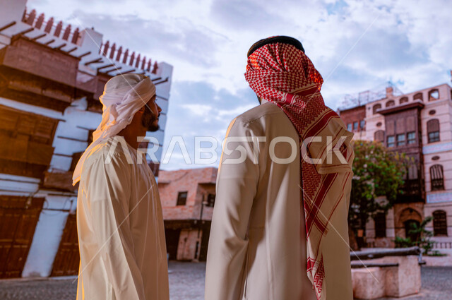 النظر الى المباني الأثرية و الأحياء الشعبيه في جده التاريخيه, الرواشين الخشبيه في البناء المعماري العربي القديم, أماكن ومعالم سياحيه أثرية مشهوره في حي البلد, رجل عربي اماراتي خليجي يرتدي العمامة والكندورة مع صديقه السعودي ويتبادلان الأحاديث والنقاشات