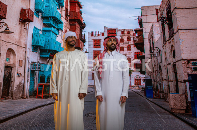 قدوم السياح من شتى أنحاء العالم، أماكن ومعالم سياحيه مشهورة في حي البلد, الرواشين الخشبيه في البناء المعماري العربي القديم, سائح عربي اماراتي خليجي يرتدي الكندورة والعمامة مع رجل سعودي يتجولان بين المباني الأثرية والأحياء الشعبيه في جده التاريخيه