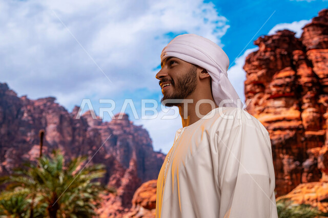زراعة النباتات و أشجار النخيل في الأماكن السياحيه المشهوره في السعوديه, الوقوف أمام التكوينات والتشكيلات الصخرية في مدينة العلا ,صورة مقربة من الجانب لسائح عربي اماراتي خليجي يرتدي الزي التقليدي يستمتع في المناظر الطبيعه الجبليه التاريخيه الأثرية العريقة