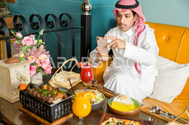 رجل عربي خليجي سعودي يجلس على مائدة الطعام و يستخدم الهاتف المحمول لالتقاط الصور  ، قضاء اجواء رائعة  في المطعم ، تشكيلة متنوعة من أصناف الطعام و الماكولات الشهية على الطاولة ، جلسات فريدة و مميزة ، مطاعم و كافيهات في السعودية