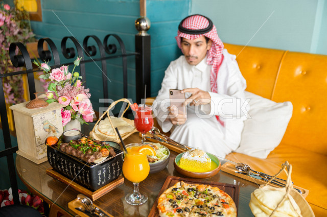 رجل عربي خليجي سعودي يجلس على مائدة الطعام و يستخدم الهاتف المحمول لالتقاط الصور  ، قضاء اجواء رائعة  في المطعم ، تشكيلة متنوعة من أصناف الطعام و الماكولات الشهية على الطاولة ، جلسات فريدة و مميزة ، مطاعم و كافيهات في السعودية