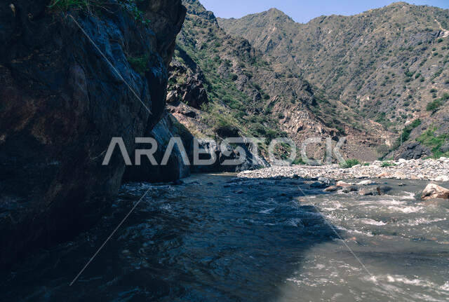 The flow of water between the valleys and mountains in the Jizan region, the mountainous environment and the green trees in the Kingdom of Saudi Arabia, the natural tourist attractions of Jizan, the peaks, heights and rock formations in Saudi Arabia.