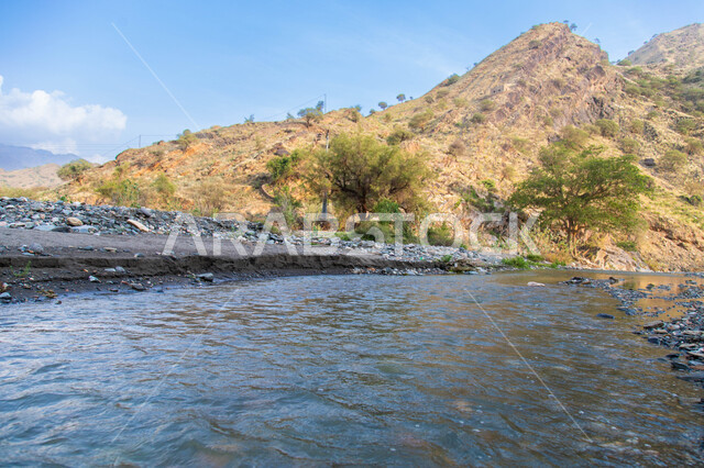 The flow of water between the valleys and mountains in the Jizan region, the mountainous environment and the green trees in the Kingdom of Saudi Arabia, the natural tourist attractions of Jizan, the peaks, heights and rock formations in Saudi Arabia.