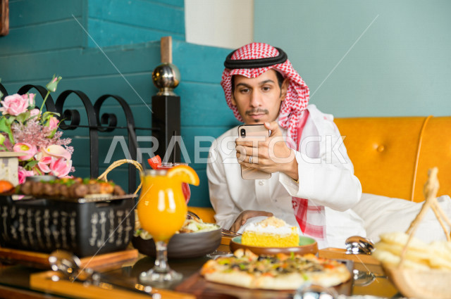 رجل عربي خليجي سعودي يجلس على مائدة الطعام و يستخدم الهاتف المحمول لالتقاط الصور  ، قضاء اجواء رائعة  في المطعم ، تشكيلة متنوعة من أصناف الطعام و الماكولات الشهية على الطاولة ، جلسات فريدة و مميزة ، مطاعم و كافيهات في السعودية